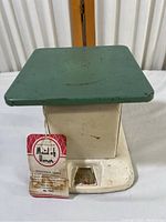 Front view of vintage Maid of Honor household scale with green square platform top and off-white base. Shows wear and marks from aging. Attached original tag visible in front.