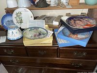 Seven collector's plates with some in boxes and papers, various artistic styles including Asian, Edna Hibel, Norman Rockwell, and floral designs.