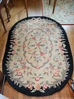 Oval hand-hooked rug with floral design and black scalloped border, laid on wooden floor.