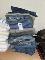 Stack of folded pants, mostly denim jeans with some tan colored pants, showing worn and frayed fabric.