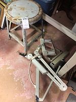 Photo of two magnifying lights with adjustable arms placed on the floor and a vintage metal stool with rusted round seat and three legs nearby.
