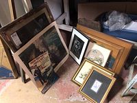 Lot of framed prints and paintings stacked upright and leaning against wall on floor, showing variety of frame styles and artwork including naval recruitment poster.