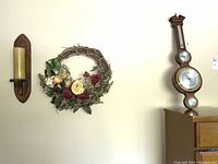 Wide shot of wall items consisting of a wooden sconce with candle, floral wreath, and Howard Miller barometer mounted on the wall.