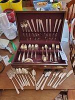 Open silverplate flatware case showcasing forks, spoons, serving pieces organized in slots and additional pieces laid out on table.