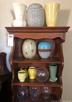Wide shot showing four shelves filled with eleven art glass and ceramic vases in a variety of shapes, colors, and finishes.