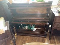 Photo of vintage two-tier wooden barrister bookcase, showing glass front open revealing interior shelves with some glassware inside. Shows condition of wood with visible wear and scratches.