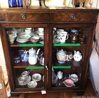 Front view of vintage wooden cabinet with glass doors showing ceramic and porcelain tea sets inside.