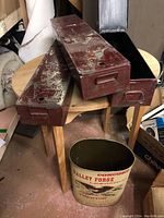 Photo shows the three red metal storage containers with hinged lids placed on the wood table. The vintage beige enamelware Valley Forge waste basket is in front of the table on the floor.