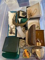 Multiple costume jewelry pieces displayed in a plastic bin including pearl-like bead necklaces, metal cuff links, a heart-shaped pendant necklace, bracelets, some in boxes and cases.