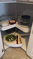 Corner kitchen cabinet with two white lazy susan shelves; upper shelf contains Corningware dishes, plastic bowls with lids, and clear glass bakeware. Lower shelf has a green ceramic bowl and chopsticks.