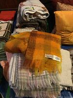 Multiple folded blankets and throws on table, including a folded orange and yellow checkered mohair throw with label visible