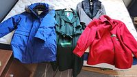 Photo showing all four outerwear items laid on a bed: blue winter coat, green overcoat, red coat, and grey blazer with faux fur collar.
