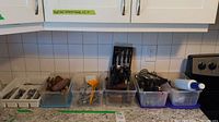 Assortment of cutlery and kitchen utensils organized in plastic bins on a countertop. Includes wooden knife block with knives and various tools like potato masher and whisks.