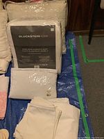 Photo showing new queen duvet packaged in plastic with Gluckstein Home label, twin polyester duvet, mattress pad and white bedding items folded on blue tarp on floor.