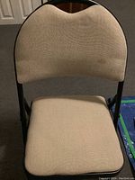 Front view of a beige fabric upholstered metal folding chair with curved backrest and padded seat, showing some wear and stain on fabric.