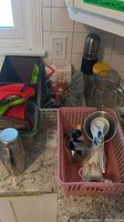 Photo showing a variety of kitchen utensils including oven mitts, measuring spoons, a cocktail shaker, and a vegetable steamer basket arranged on a counter with bins