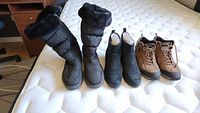 Three pairs of ladies boots: tall black winter boots with fur trim, black suede ankle boots, and brown hiking boots laid out on mattress.