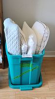 Teal plastic bin filled with three white bed pillows and one quilted back rest pillow.