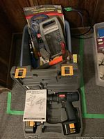 Ryobi cordless drill in open carrying case with user manual on top. Bin with multiple hand tools and digital multimeter behind case.