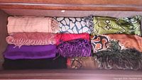 Nine assorted shawls in various colors and patterns laid flat in a drawer.