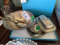Photo showing a large silver plate serving bowl with fork and spoon inside, and a separate silver plate butter dish with a rectangular lid and decorative edge on a blue box.