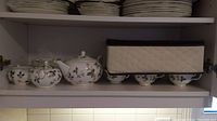 Lower cabinet shelf showing sugar bowl, creamer, teapot and fabric storage container on wooden rack