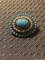 Photo showing an oval antique brooch with a central turquoise stone, surrounded by small pearls and turquoise beads on a textured surface.