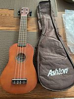 Full view of Ashton soprano ukulele with nylon strings and natural wood finish beside a soft brown carrying case with Ashton logo.
