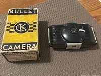 Photo showing the black Bullet Camera and its original yellow and black Eastman Kodak box on a brown surface.