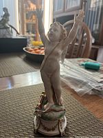 Front view of the cherub figurine showing arms raised and facial details with natural colors and gold-accented pedestal base.