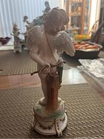 Rear view of the porcelain cherub figurine showing wings and bow, standing on painted base with green and gold accents.