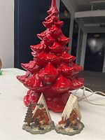 Red ceramic Christmas tree lamp base along with two small Christmas scene figurines with snowy rooftops, placed on a table with a dark background.