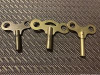Photo of three metal clock keys: a small, medium, and large key with double-hole winders on a dark textured surface.