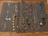 Top-down image of 13 pieces of costume jewelry laid out on a table, including various chains, necklaces, bracelets, and pendants of gold and silver tones, faux pearls, beads, and cord materials.