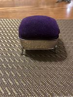 Closed ring box showing purple velvet cushion on top and sterling silver engraved base with four feet.