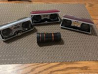 Four pairs of folding opera glasses arranged around a black monocular telescope on a woven placemat and wooden table.