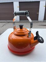 Side view of bright orange enamel Le Creuset kettle with black handle and whistling spout cover on white table.