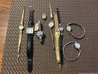 Photo of nine women's wristwatches laid out on a textured surface showing various styles and band materials including metal links, leather, and stretch bands.