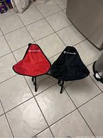 Photo showing two triangular foldable camp stools placed side by side on a tiled floor. One has a red fabric seat, the other black. Both seats have the Outbound logo in white.