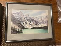 First framed painting showing a mountain lake with snowy cliffs, turquoise water, and pine trees, signed S. Sadler 1983.