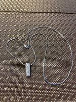 Full view of the silver necklace with rectangular pendant and clasp laid out on textured surface.