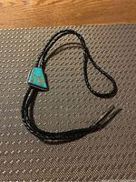Full view of the bolo tie showing the braided leather cords, large turquoise stone centerpiece, and metal tips.