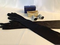 Pair of long black vintage gloves, mother of pearl opera glasses, and cream textured case arranged on white background.