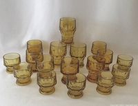 Front view showing 17 amber glass tumblers arranged in groups by size and color tone on a white fabric background