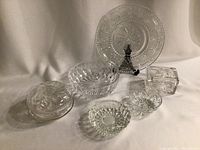 Lot of six vintage crystal and glass items on white fabric background including bowls, dishes, trinket box, and cake plate.