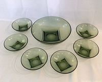 Seven green translucent glass bowls arranged on white surface, one large salad bowl and six smaller matching bowls, all with square embossed design at center.