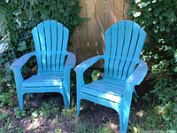 Pair of turquoise plastic Adirondack chairs outside by a wooden fence with green plants in the background, showing overall condition and color.