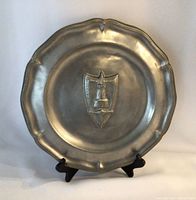 Full view of the vintage pewter plate on a black stand showing scalloped edge and embossed emblem in the center.