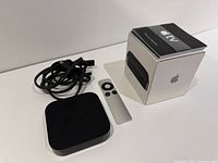 Apple TV unit, remote control, power cable, manual, and box arranged on white surface.