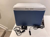 Front view of blue and white Coleman cooler with 12V car power adapter and 110V wall power adapter cords coiled beside it.